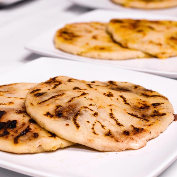 Close-up of two golden-brown pupusas with dark grill marks on a white plate, with more in the background.